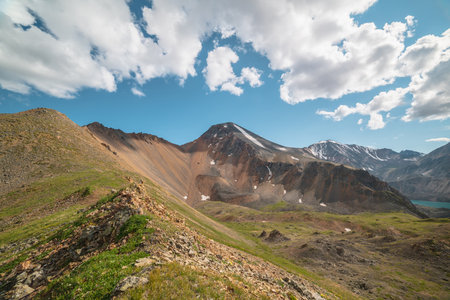 Scenic landscape with sunlit green grassy stone hill and high orange mountain ridge under white clouds in blue sky. Colorful scenery with rocky mountains and sharp rocks in sunlight under cloudy sky.の写真素材