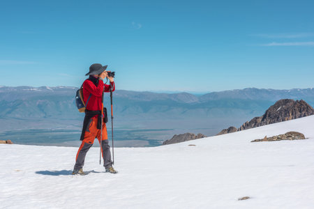 Tourist in red walks on snow mountain near abyss edge on high altitude under blue sky in sunny day. Man with camera on snowy mountain near precipice edge with view to large mountain range in away.の写真素材