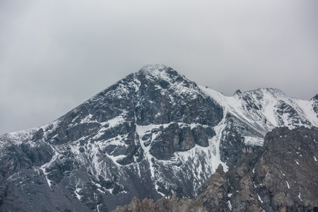 Awesome landscape with high snowy mountain peaked top with sharp rocks in cloudy sky. Dramatic view to snow mountain pointed peak in rainy weather. Atmospheric scenery with white snow on black rocks.の写真素材