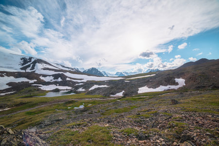 Colorful landscape with mountain lake among hills and rocks against sunlit high snowy mountain range under cloudy sky in changeable weather. Awesome sunny scenery with lake and snowy mountain top.の写真素材