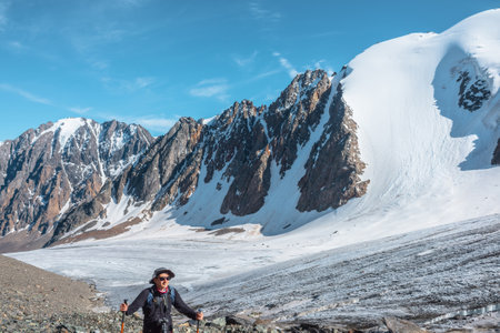 Hiker with trekking poles walks near large glacier tongue in high mountains. Scenic landscape with tourist near glacier in sunlight. Man with sunglasses and camera among snow mountains in sunny day.の写真素材