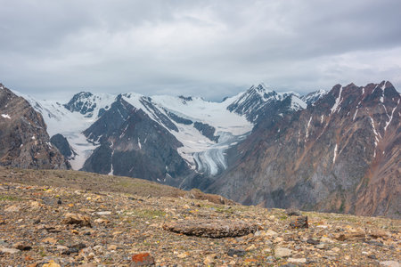 Dramatic landscape with high snowy mountain range with long glacier tongue and pointed peak in sunlight under cloudy sky. Atmospheric view from stone hill to large snow mountains at changeable weatherの写真素材
