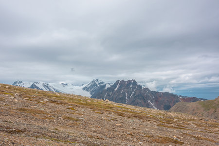 Dramatic landscape with sunlit diagonal stone mountainside with view to high snow mountains with sharp rocks and pointed peak under cloudy sky. Beautiful scenery with peaked top at changeable weather.の写真素材