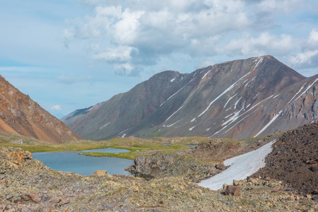 Colorful landscape with beautiful lake in sunlit mountain valley among rocks and high mountain ridge under cloudy sky in changeable weather. Awesome scenery with alpine lake among stones in sunlight.の写真素材