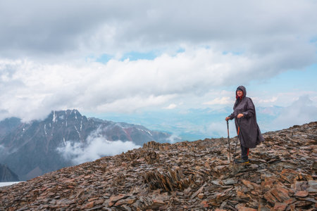 Scenic alpine landscape with tourist on stone hill with view to snow mountain peak under cloudy sky. Hiker with trekking poles at very high altitude with view to snowy mountain top and low clouds.の写真素材