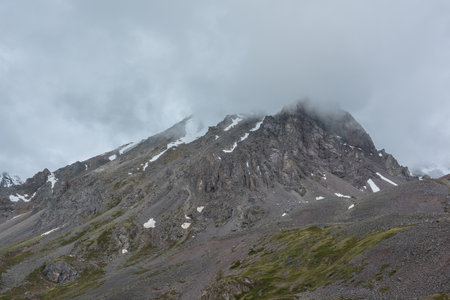 Dramatic mountain landscape with sharp rocks with snow in rainy low clouds. Beautiful scenery with large rocky mountain with snow top in low gray cloudy sky. Gloomy rainy weather in high mountains.の写真素材