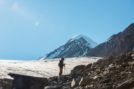 Scenic alpine landscape with silhouette of hiker with trekking poles against large glacier tongue and snow mountain peak in sunlight. Man with backpack in high mountains under blue sky in sunny day.の写真素材