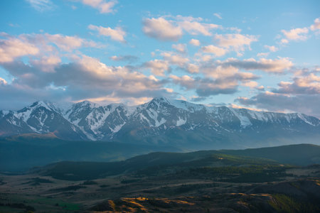 Beautiful aerial view to sunlit golden forest hills against high snowy mountain range in sunlight under cloudy sky in sunset colors. Colorful landscape with large snow mountains in gold sunshine.の写真素材