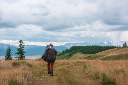 Tourist walks through hills and forest towards bad weather. Hiker on way to large snow mountain range under rainy cloudy sky. Man in raincoat in mountains in overcast. Traveler goes towards adventure.の写真素材