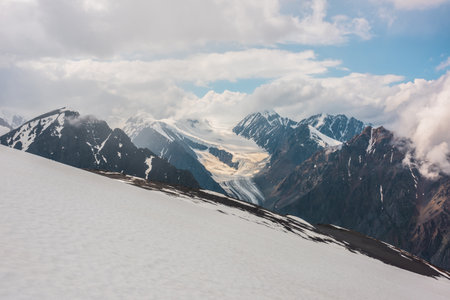 Dramatic landscape with long glacier tongue and high snow mountains in cloudy sky. Wonderful aerial view to sunlit large glacier and snowy mountain peak in low clouds. Top view to majestic mountains.の写真素材