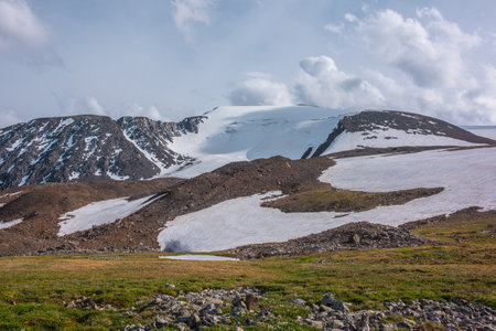 Colorful green landscape with shadow from cloud on sunlit snowy mountain top under cloudy sky in changeable weather. Awesome sunny scenery with lake in grass meadow against high snow mountain range.の写真素材