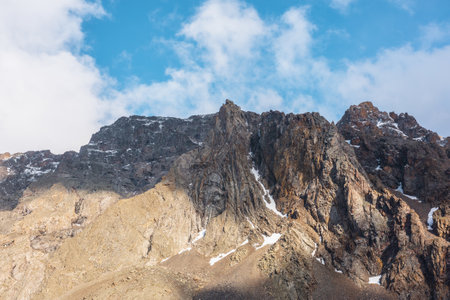 Atmospheric landscape with sharp pinnacle on sunlit high mountain range under clouds in blue sky. Beautiful alpine scenery with peaked mountain top in sunlight under cloudy sky at changeable weather.の写真素材