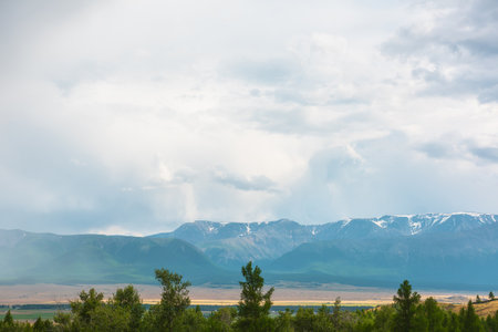 Dramatic view from forest to high mountain range in sunlight during rain in changeable weather. Colorful landscape with green forest and sunlit steppe against large mountains under cloudy sky in rain.の写真素材