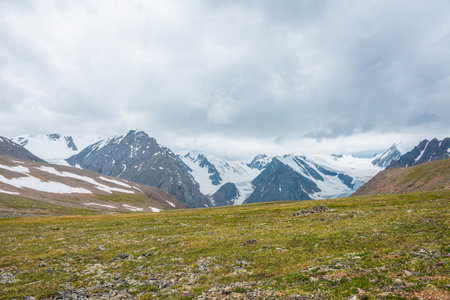 Scenic alpine view from sunlit green grassy hill to high snowy mountain range with sharp tops and glaciers under gray cloudy sky. Colorful landscape with large snow mountains at changeable weather.の写真素材
