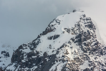 Dramatic landscape with sunlit high snowy mountain top in low cloudy sky. White snow on black rocks with golden shine in low clouds. Awesome snow mountain peak with gold sunlight in changeable weatherの写真素材
