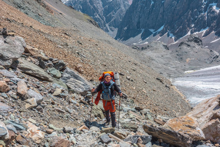 Backpacker with trekking poles walks in high mountains in sunny day. Scenic alpine landscape with tourist near glacier in sunlight. Man with sunglasses and camera among mountains with snow in sunshineの写真素材