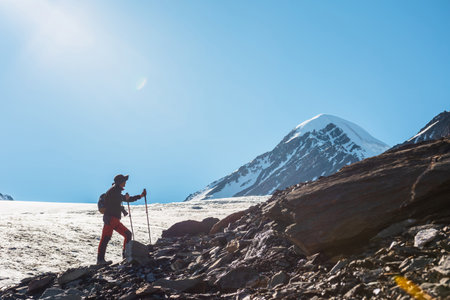 Scenic alpine landscape with silhouette of hiker with trekking poles against large glacier tongue and snow mountain peak in sunlight. Man with backpack in high mountains under blue sky in sunny day.の写真素材
