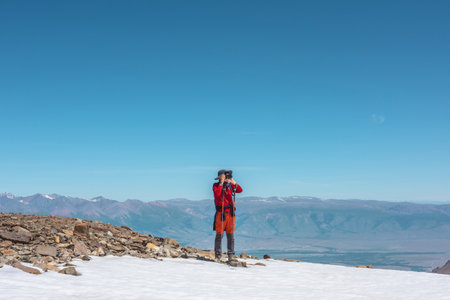 Tourist in red walks on snow mountain near abyss edge on high altitude under blue sky in sunny day. Man with camera on snowy mountain near precipice edge with view to large mountain range in away.の写真素材