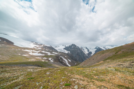 Scenic panoramic view from sunlit green grassy hill to high snowy mountain range with sharp tops and glaciers under gray cloudy sky. Colorful landscape with large snow mountains at changeable weather.の写真素材