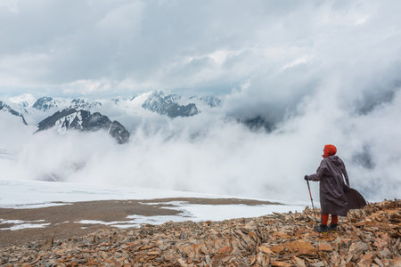 Hiker and high snowy mountains in dense low clouds. Minimal scenery with man on stone hill against beautiful mountain peaks in thick clouds. Simple minimalism with man and majestic mountains nature.の写真素材