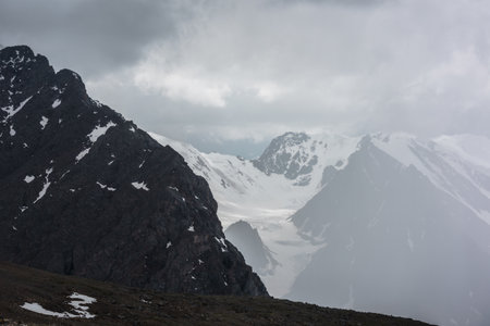 Atmospheric landscape with fuzzy silhouettes of sharp rocks and snowy mountains in low clouds during rain. Dramatic view to large snow mountains and glacier blurred in rain haze in gray low clouds.の写真素材