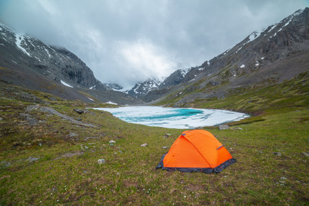 Atmospheric mountain landscape with orange tent near frozen alpine lake and snowy mountains. Awesome scenery with icy mountain lake on background of snow mountains in low clouds. Tent near ice lake.の写真素材