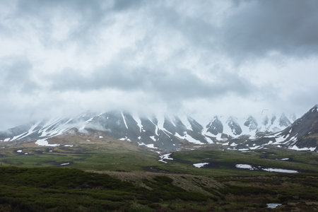 Dramatic alpine landscape with snowy mountains in gray low clouds. Bleak atmospheric scenery of tundra under lead gray sky. Gloomy minimalist view to mountain range among low rainy clouds in overcast.の写真素材