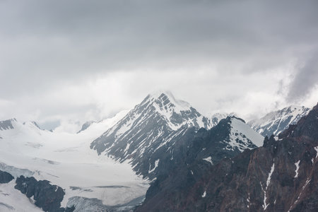 Atmospheric landscape with sharp rocks and high snowy mountain top in rainy low clouds at overcast. Dramatic gloomy scenery with large snow mountains and glacier in gray cloudy sky at rainy weather.の写真素材
