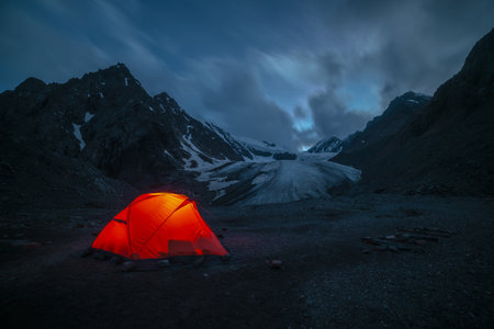Awesome mountain landscape with vivid orange tent near large glacier tongue under clouds in night starry sky. Tent glow by orange light with view to glacier and mountains silhouettes in starry night.の写真素材