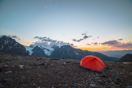 Scenic alpine landscape with tent at very high altitude with view to large mountains in orange dawn sky. Vivid orange tent with awesome view to high mountain range under cloudy sky in sunset colors.の写真素材