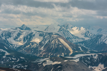 Awesome aerial view to high snow mountain peaks in sunlight in overcast. Atmospheric mountain landscape at high altitude with cloudiness. Alpine view to snowy mountain tops in light under cloudy sky.の写真素材