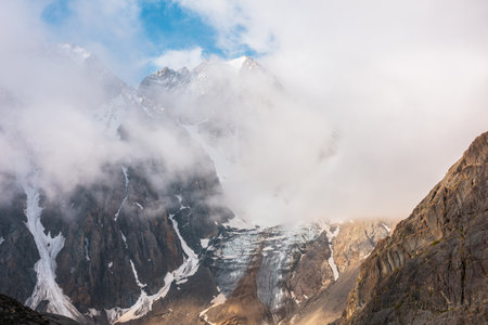 Scenic mountain landscape with snowy mountain top and glacier in dense low clouds in sunrise colors. Colorful mountain view to vertical glacier with icefall in morning sunlight in thick low clouds.の写真素材