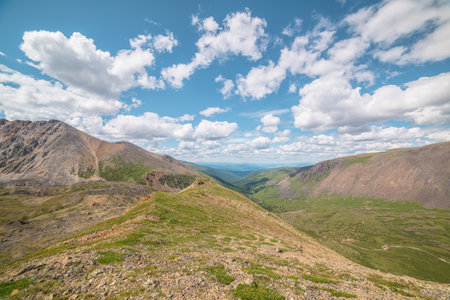 Scenic view from mountain ridge to green forest valley among mountain ranges and hills on horizon at changeable weather. Green landscape with sunlit mountain vastness under cumulus clouds in blue sky.の写真素材