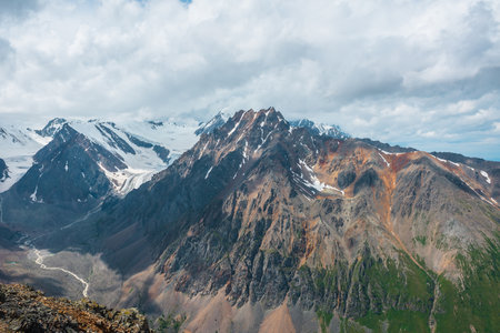 Vivid mountain landscape with sunlit large pinnacle with snow under cloudy sky. Picturesque scenery with high mountain peak with shadows from clouds at changeable weather. Sharp rocks in sunlight.の写真素材