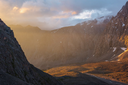 Scenic alpine landscape with mountain wall in low clouds in golden sunrise colors. Colorful view to high mountains and rocks in gold morning sunlight. Golden light in mountain valley at early morning.の写真素材