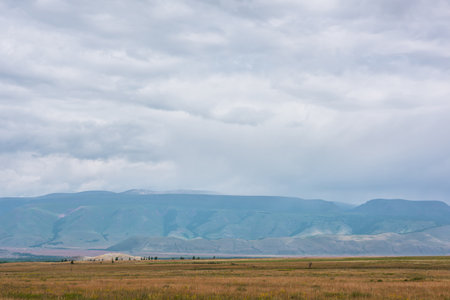 Dramatic alpine view to sunlit steppe and somber large mountains in low clouds during rain. Gloomy mountain landscape with bleak mountain range in rain and steppe in sunlight in changeable weather.の写真素材