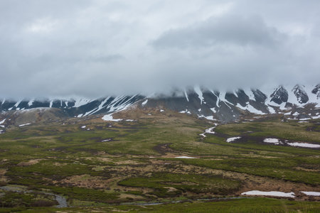 Dramatic alpine landscape with snowy mountains in gray low clouds. Bleak atmospheric scenery of tundra under lead gray sky. Gloomy minimalist view to mountain range among low rainy clouds in overcast.の写真素材