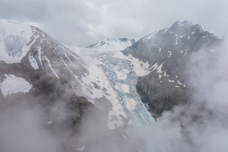 Atmospheric mountain landscape with long vertical glacier tongue with cracks among rocks in dense low clouds. Awesome aerial view to large glacier with icefall in thick low clouds in high mountains.の写真素材