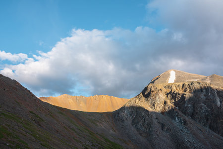 Scenic landscape with sunlit wide sharp mountain ridge between mountain tops at changeable weather. Colorful mountain scenery with large sharp rocks on ridge top in sunlight under clouds in blue sky.の写真素材