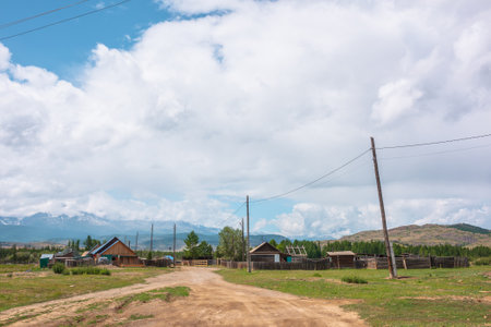 Dramatic alpine landscape of beautiful mountain village with wooden rustic houses with sloping roofs in sunlight and large mountain range in low clouds. Russia, Altai Republic, Kurai, 13 July, 2021.のeditorial素材