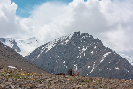 Small cabin at high altitude near precipice edge with view to high rocky mountain with peaked top. Small house against snowy pointy peak under cloudy sky. Russia, Altai Republic, Aktru, 06 July, 2021.のeditorial素材