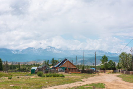 Dramatic alpine landscape of beautiful mountain village with wooden rustic houses with sloping roofs in sunlight and large mountain range in low clouds. Russia, Altai Republic, Kurai, 13 July, 2021.のeditorial素材