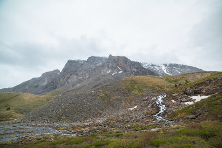 Atmospheric landscape with clear mountain stream from hills against snowy mountain range with peaked top in low clouds. Beautiful mountain creek and high sharp rocks under cloudy sky in gloomy weatherの写真素材