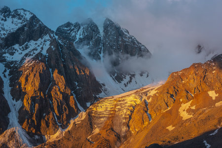 Scenic mountain landscape with snowy mountain top and glacier in low clouds in golden sunrise colors. Colorful mountain view to vertical glacier with icefall in gold morning sunlight in low clouds.の写真素材