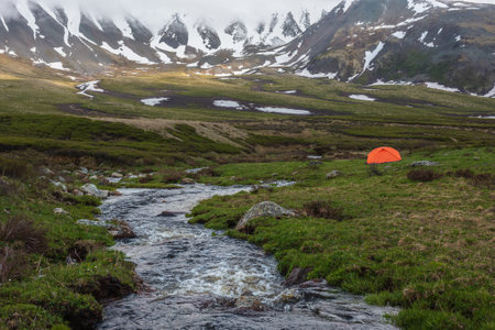 Orange tent near clear water stream with view to mountain range among low rainy clouds. Dramatic alpine landscape with mountain creek and snowy mountains in gray low clouds. Gloomy scenery of tundra.の写真素材