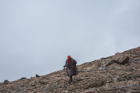 Tourist on stone hill at very high altitude under gray cloudy sky. Hiker with trekking poles and camera on rocky mountain in overcast weather. Man in high mountains among sharp rocks and sharp stones.の写真素材