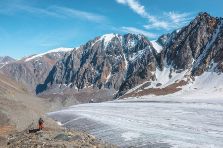 Hiker with trekking poles near large glacier tongue in high mountains. Scenic alpine landscape with tourist near glacier in sunlight. Man with sunglasses and camera among snow mountains in sunny day.の写真素材