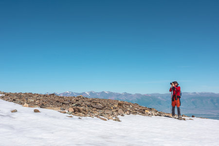 Tourist in red walks on snow mountain near abyss edge on high altitude under blue sky in sunny day. Man with camera on snowy mountain near precipice edge with view to large mountain range in away.の写真素材