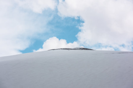 Beautiful landscape with huge snowy mountain dome with rocky top under big lush white clouds in blue sky. Awesome mountain scenery with high snow mountain in dome shape in center at changeable weatherの写真素材