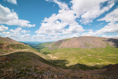 Scenic view from mountain pass to green forest valley among mountain ranges and hills on horizon at changeable weather. Green landscape with sunlit mountain vastness under cumulus clouds in blue sky.の写真素材